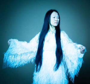 Woman with long black hair wearing a white fringed dress extends her arms, blue studio lighting.