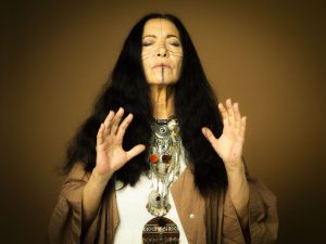Woman with long dark hair and layered silver jewelry, eyes closed, hands raised as if in meditation.