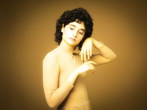 Female dancer in a beige leotard posing with arched arms against a warm golden studio backdrop.