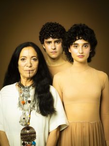 Three models pose together: an older woman with facial paint and bold tribal jewelry, flanked by a man and a woman in peach-toned outfits against a brown backdrop.