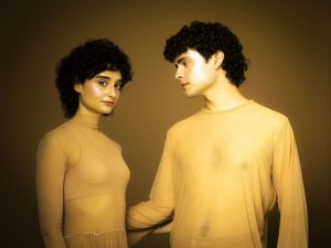 Two models with curly hair wear beige outfits and face each other in a warm brown studio setting.