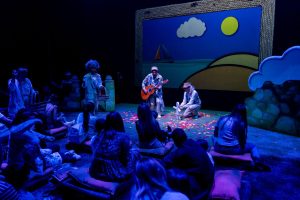 Children in a blue-lit theater watching a stage performance with musicians; a guitarist and a singer in costumes perform on a colorful set.