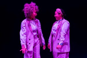 Two performers in matching lavender outfits facing each other and smiling on a dark stage with pink lighting.