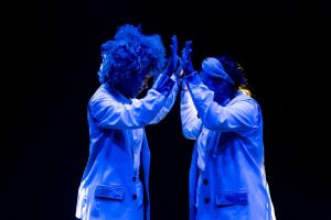 Two people in white lab coats high-five on a dark stage, illuminated by blue light.