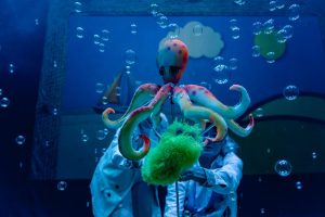 Colorful octopus puppet on a rod held by two people in lab coats underwater, bubbles around it.