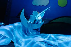 Blue-lit theatrical scene showing a round-headed figure on a crescent moon prop, with flowing fabric around them.