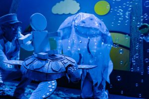 Blue-toned scene of a person in a lab outfit adjusting a large paper turtle model in an underwater-themed display, with bubble props around it.