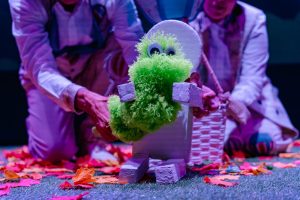 Bright green fuzzy puppet with googly eyes sits on a small block-built figure, being positioned by two people in light clothing on a leaf-strewn lawn.