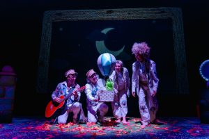 Four performers in white patchwork costumes on a dark stage, passing a wicker basket with a blue striped balloon inside.