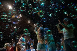 Group of children at a nighttime celebration reaching for colorful bubbles and confetti lights above them, joyful and playful scene
