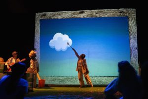 Performer on stage raises a wand to a white cloud prop against a blue gradient backdrop, framed by a textured border box on stage.