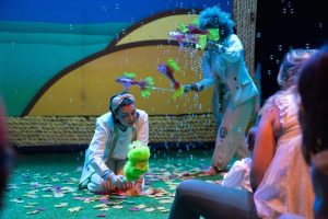 Two performers in white costumes entertain a child audience; one kneels with a bright green fuzzy frog puppet as bubbles float nearby.
