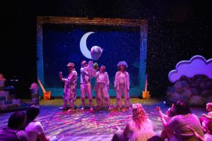 Stage show for kids: four performers in white costumes on a colorful night-sky set with a crescent moon, while children sit on the floor watching.