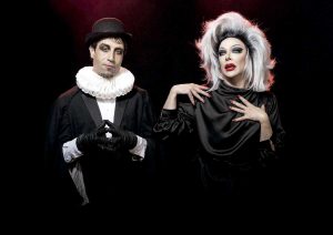 Two performers pose against a dark background: a mime in a black suit with a white ruff and bowler hat, hands in a triangle, and a drag performer with a platinum wig and dramatic makeup.