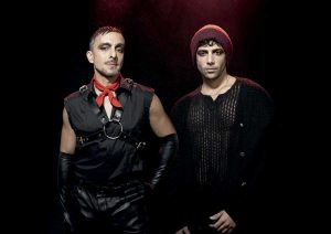 Two men in cosplay posing against a black background; one in a black leather harness with a red scarf, the other in a black knit top and red beanie.