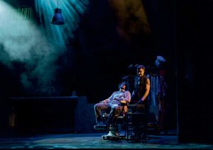 Two performers on a dark theater stage under blue spotlight; one sits in a rolling chair, the other stands beside a clothing rack amid smoke.