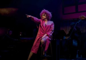Drag performer in a pink satin robe with bright pink hair points toward the audience on a dim stage.