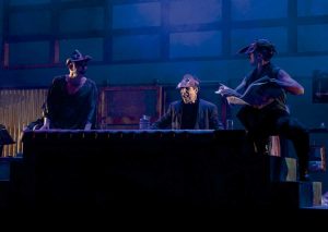 Three performers on a blue-lit stage gathered around a long black table; the center actor wears a headlamp and laughs.