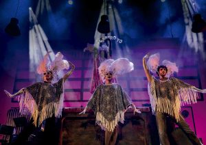Three dancers in glittery fringe outfits with pink feather headpieces perform on a colorful stage under dramatic spotlights.