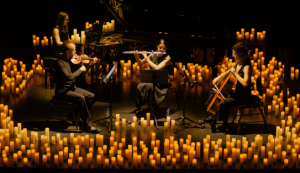A quintet of classical musicians performing around a grand piano, surrounded by dozens of lit candles in a dark stage.